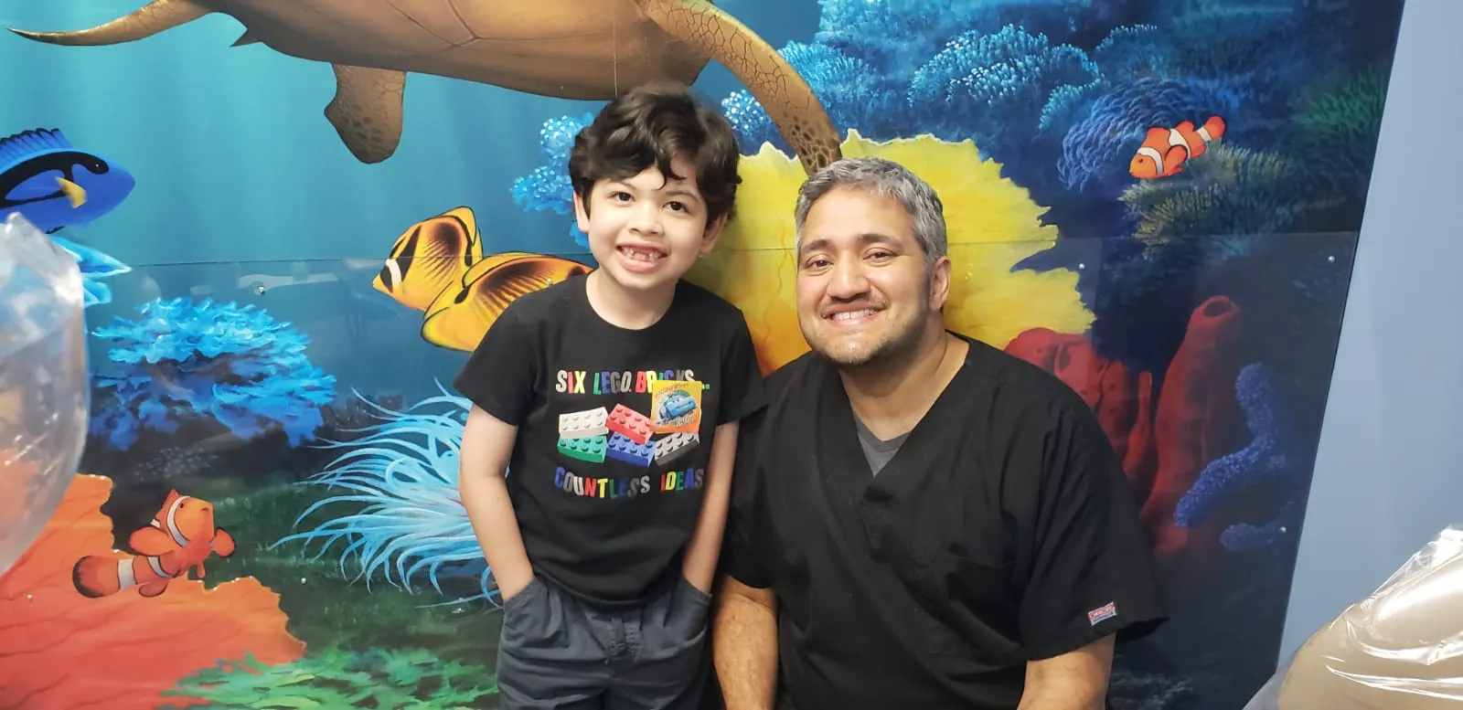 Smiling boy with his dentist in front of a colorful ocean mural at Dental Care Kids